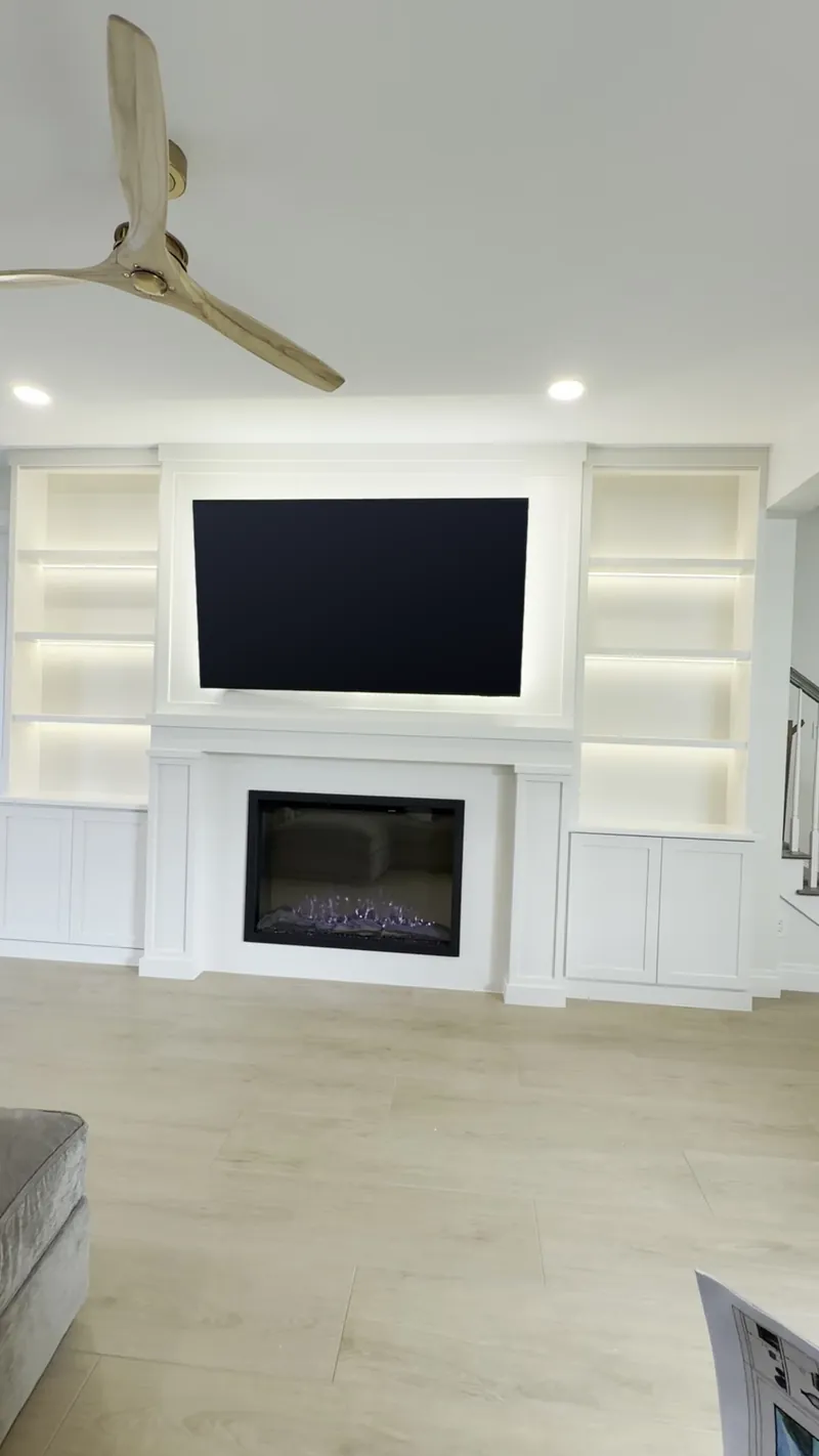 All-white built-in wall with a classic fireplace surround, a backlit TV panel and tall lit towers over Shaker cabinets